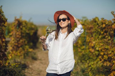 Güneş gözlüğü ve şapka takan kadın şarap garsonunun elinde şarap kadehini tutuyor ve onu tadıyor..