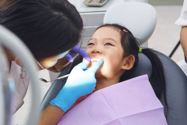 Dişçi koltuğunda oturan kız çocuğu dişçiden rutin kontroller alıyor. Diş hekimi dişlerini ve ağız sağlığını dikkatlice kontrol ediyor. Her şeyin iyi durumda olduğundan emin oluyor..