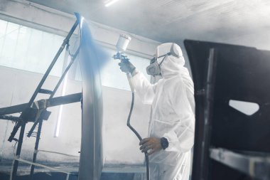 Erkek endüstriyel işçiler, arabanın koruyucu maske ve atölye garajında giyilen kıyafetlerin bir kısmına hava fırçası tabancası kullanarak boya sıkarlar. Adam kaputa boya püskürtüyor.. 