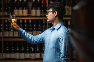 Şarap mahzeninde dikilen odaklanmış Asyalı şarap garsonu, elinde beyaz şarap kadehiyle, rengini ve berraklığını ölçmek için, çeşitli şarap şişeleriyle dolu rafların önünde duruyor..