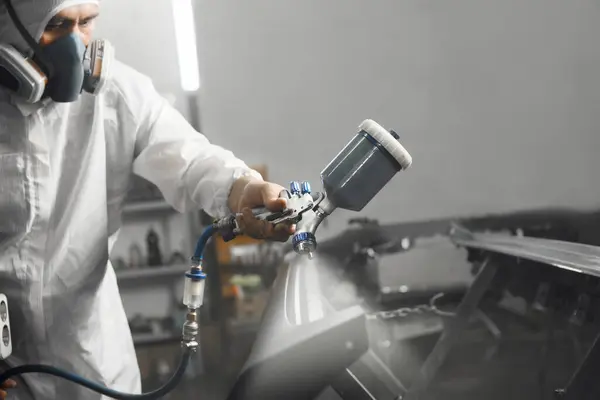 Maskeli otomobil tamircisinin ve koruyucu iş giysisinin yakın plan çekimi boya odasındaki sprey tabancayla arabaya uygulanır. Kazadan sonra aracın restorasyonu. 