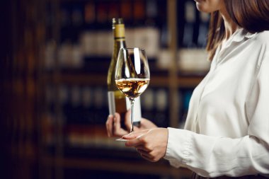 Kapalı alanda yüzü olmayan, beyaz gömlekli, el ele tutuşma şişesi ve mahzene dökülen beyaz şaraplı kadın portresi..