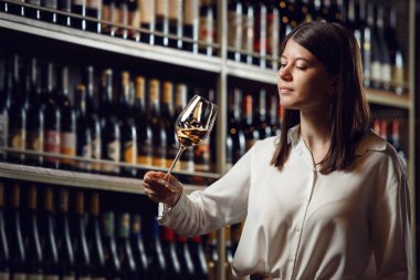 Şarap tüccarı kadın mahzendeki bardaktan beyaz şarap tadıyor. Şarap kadehini arka planda tutan kadın şarap garsonunun yakın çekimi. Şarap yapma kursları, şarap tatma. Sommelier Birliği