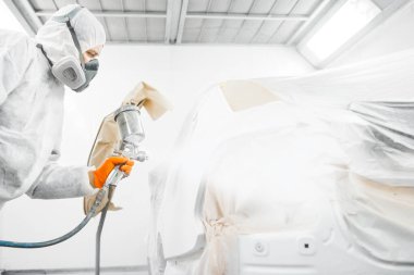 Tamirat sırasında araba tamircisi boya odasına sprey tabancayla beyaz bir araba boyuyor. Arabanın onarımı için servis.