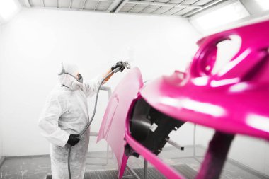 Koruyucu maskeli ve elbiseli bir adam boya kabinindeki sprey tabancayla araba elementlerine cila sıkar. Otomobili boyadıktan sonraki kaplama işlemi.