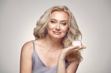 Stüdyo portresi, gri izole edilmiş arka planda pijamalı, sağlıklı bir ten rengine sahip nemlendirici tutan olgun sarışın. Yaşlıların ciltlerinin tazelenmesi kavramı.