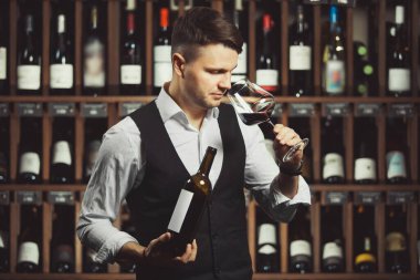 Elinde şişe ve bir bardak kırmızı şarap tutan, yaşını belirtmek için kokusunu alan adam, ürün hakkında bilgi yazmaya hazır, bulanık arka planda şarap rafı olan mahzen.