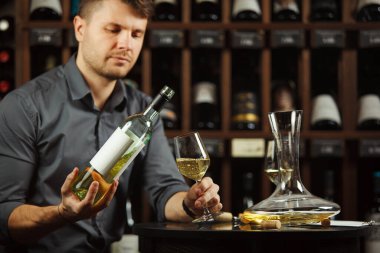 Sommelier erkeği şişeden beyaz şarap dolduruyor. Açıya etiketi yapıştırıyor. Alkollü içecek israfı, kaplar ve içeceklerle dolu mahzen.