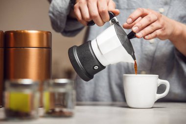 Person pours stream of freshly brewed coffee from stovetop Moka pot into small white cup, close up photo. clipart