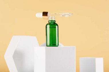 Boynunda pipet kapağı olan açık yeşil damlacıklı mini şişe, sarı arka planda beyaz geometrik şekillerle çevrili küp üzerinde duruyor. Uzayı kopyala Güzellik ürünleri pankartı, şablon.