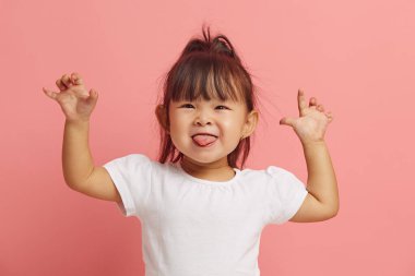 Küçük güzel Asyalı kızın stüdyo portresi eğleniyor ve ellerini pembe izole edilmiş arka planda kaldırarak dilini gösteriyor..