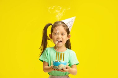 Koreli güzel kız çocuğunun münferit bir resmi, beş yaşında gibi görünüyor, parti şapkası takıyor, doğum günü pastasının üzerindeki mumları söndürüyor, dilek tuttuktan sonra neşeli ifade ediyor.