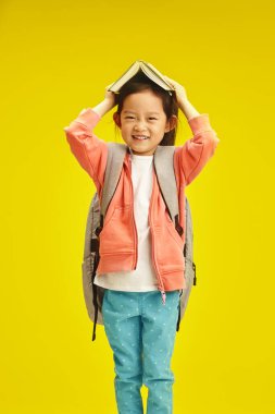 Gülümseyen Asyalı etnik kökenli bir kız öğrencinin dikey çekimi omuzlarında sırt çantasıyla sarı izole altyapının üzerinde duran açılış kitabını kafasında taşıyor. Koreli küçük kız hazırlanıyor