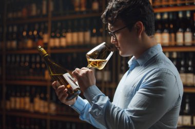 Professional sommelier, wearing glasses and light blue shirt, carefully examines glass of white wine by smell it, while attentively reading label on wine bottle standing against background of wine clipart