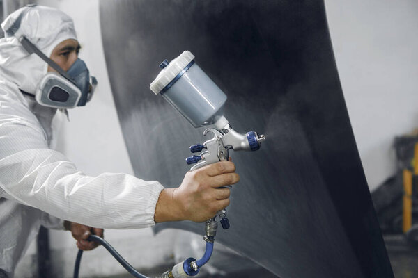 Spray painter worker in protective glove with airbrush pulverizer varnishing and painting car body in paint chamber. Car vehicle part in garage during repair work.