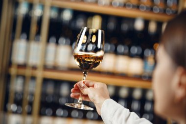 Kadın barmenin kapalı mekanda çekilmiş fotoğrafı beyaz şarabı derecelendirir, rengine bakar, alkolik bir içkinin asitliğini ölçer, farklı şaraplarla dolu raflarda durur. Sommelier okulu, içecekler