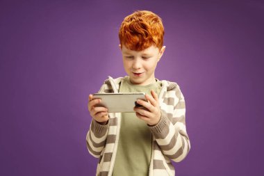 Studio portrait of red haired little boy passionate playing game on smartphone, unable to break away from the game, wearing in casual clothes, standing against violet isolated background. Dependence clipart