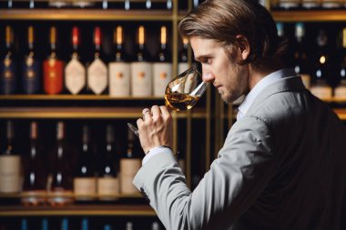 Profile photo of european ethnicity male sommelier appreciating white wine from glass by smelling standing in cellar against fulled wooden shelf background with bottles. Man tasting alcoholic drink in clipart