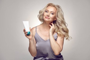 Stüdyoda, elinde beyaz kozmetik krem tüpü tutan yaşlı ve memnun bir kadının resmi var. Gri izole arka planda bedava kopyasal alan var. Yaşlı cilt ve cilt bakımının yenilenmesi.