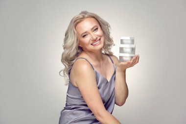 Cilt bakımı için yüzünün yanında birkaç kavanoz krem tutan lüks, olgun bir kadın. Evde pijama giyen yaşlı sarışın kadın gri bir arka planda sırıtan çeşitli nemlendiriciler ve zevkler taşıyor..