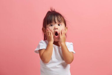 Şaşırmış üç yaşındaki küçük kızın stüdyo portresi ağzını açık tutuyor, elleri yanaklarında, şokla kameraya bakıyor, pembe arka planda izole edilmiş. Çocuk çok şaşırdı.. 