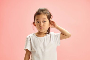 Studio indoor portrait of surprised Asian ethnicity child girl try to overhear listening intently, wearing in neutral white t shirt isolated on soft pink background with a free copy space. Curiosity clipart