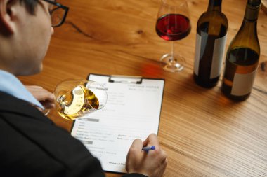 Odaklanmış şarap garsonunun bodrumda elinde bir bardak şarap ve pano tutarken, şarap tatma oturumuna hazırlanırken, şarapların özelliklerini analiz ederken resmedilmiş..