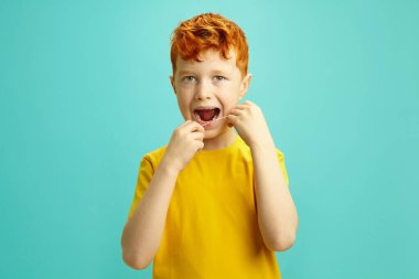 Stüdyo portresi, diş ipi kullanan, ağzı açık diş ipi kullanan, mavi arka planda belden izole edilmiş, bedava fotokopi alanı olan kızıl saçlı bir çocuk. Çocuk ağız temizliği konsepti. Çocukların dişi
