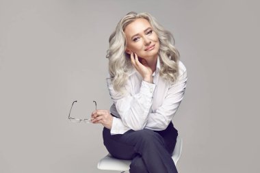 Elli yaşında, oldukça yaşlı bir kadının lüks bir kadın portresi. Sandalyeye oturmuş gri izole edilmiş, şık günlük kıyafetler giymiş, gözlük takmış, sana bakıyor..