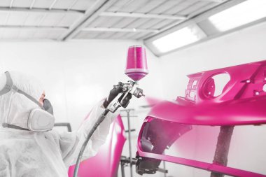 İşçi araba tamponunu sprey silahla boyuyor. Tamir sırasında boya odasındaki pembe araba parçası. Kazadan sonra aracın restorasyonu.