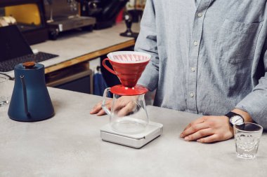 Yetenekli barista kafe tezgahında duruyor, V60 hunisi kullanarak dikkatlice kahve hazırlıyor. Dijital ölçekte tam olarak mayalandığından emin olmak için huni, sıcak suyla birlikte cam sürahinin üzerinde duruyor.