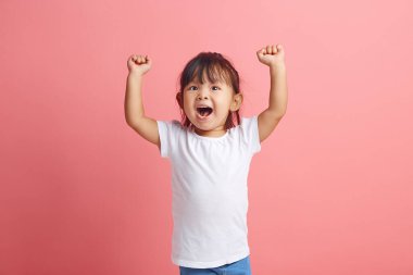 Olumlu küçük Asyalı kızın stüdyo portresi sevinçle el ele tutuşur. Kaygısız bir gülümsemesi vardır. Pembe arka planda izole edilmiş beyaz tişörtler giyer..