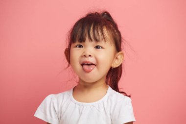 Neşeli küçük Asyalı kızın stüdyo portresi kameraya dilini gösteriyor, pembe izole bir arka planda dururken neşeli bir şekilde gülümsüyor. Neşeli çocuk portresi. 