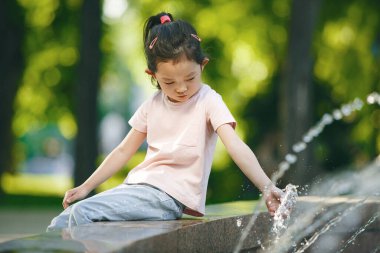 5-6 yaşlarında, pembe t-shirt ve mavi kot giyen Asyalı bir kızın açık hava portresi fıskiyenin kenarında oturuyor, elini yeşil ağaçların arka planına uzatarak suya uzanıyor.