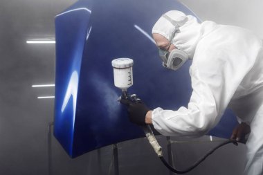 Koruyucu maske takan ve tulumlu işçiler boyalı odada boyalı boya kullandıktan sonra araba gövdesi elemanlarına cila püskürtüyorlar. Adam sprey tabancayla çalışıyor.