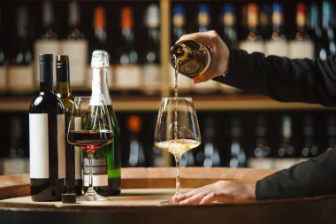 Kimliği belirsiz biri şarap dükkanının mahzeninde şişeden bardağa beyaz şarap döker, içi şarap dolu ahşap raflarla doludur.. 