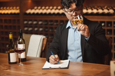 İçerideki profesyonel şarap uzmanı beyaz şarap kokusu alıyor masada oturuyor ve içki listesiyle ilgili notlar alıyor, mahzendeki elit alkollü şişelerle dolu ahşap raflara karşı..