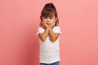 Korkmuş küçük Asyalı etnik kız kameraya korku dolu gözlerle bakıyor, bir şeylerden korkuyor, ellerini yanaklarından ayırmıyor, korkmuş görünüyor, pembe arka planda izole edilmiş. Duygusal olarak dehşete düşmüş çocuklar.