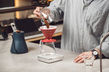 Barista 'nın yakından çekilmiş fotoğrafı, kahve çekirdeklerini titizlikle cam sürahinin üzerinde dinlenen kırmızı kahve filtresine döküyor, ki bu dijital ölçekte ideal bir kahve-su oranı için mükemmel.