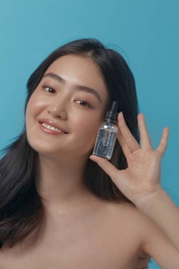 Yüzünün yanında yüz serumu tutan neşeli Japon kadın mavi bir arka planda poz vermekten zevk alıyor ve gerçek portreyi kapatıyor. Yüz bakımı, yüz bakımı, kozmetoloji
