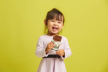 Neşeli üç yaşındaki kız neşe içinde ve zevkle lezzetli siyah çikolata yiyor ve sarı izole edilmiş arka planda komik bir şekilde duruyor..