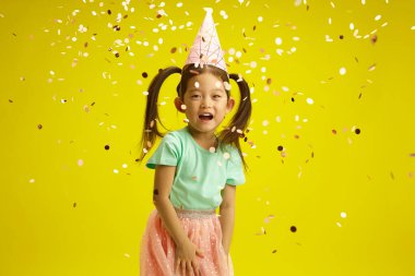 Neşeli görünümlü Koreli küçük bir çocuk koni şapkalı neşeli bir şekilde konfeti fırlatıyor, kutlama havasında, canlı sarı izole bir arka planda duruyor.
