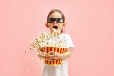 Üç boyutlu gözlük takan, elinde kırmızı patlamış mısırla farklı yönlere uçan, çizgi film ya da film başlamadan önce bir beklenti ifadesi olan mutlu küçük bir kızın stüdyo portresi.