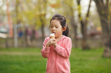 Pembe kapüşonlu çocuk, yeşil çimenleri ve ağaçları gösteren bulanık arka planda yaz günü parkta dondurma yemekten hoşlanıyor..