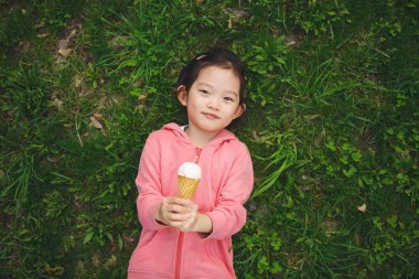 Asyalı küçük bir kız yeşil çimlerin üzerinde sırt üstü uzanmış pembe kazak giyiyor, kameraya bakıyor ve elinde beyaz dondurma külahı tutuyor.. 