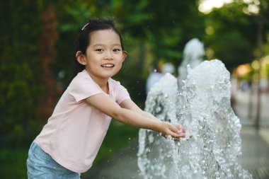 5-6 yaşlarında Asyalı bir kızın açık hava portresi. Günlük pembe tişört ve kot pantolon giyiyor. Yazın serin spreyin tadını çıkarırken çeşmede suyla gülüyor ve oynuyor.