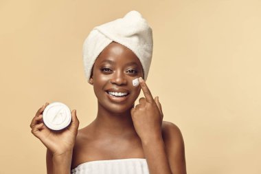 Saç türbanı takmış kahverengi tenli, vücut havlusu krem teneke tutuyormuş ve yüzüne sürüyormuş. Afrikalı Amerikalı genç bir kadın yuvarlak metal kavanozda kozmetik ürünler gösteriyor. Güzellik bakımı.