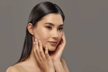 Güzel kız, güzellik tedavisinden sonra yumuşak teninin tadını çıkarırken elleriyle yüzüne dokunuyor. Gri renkli Asyalı genç bir kadın modelin güzellik stüdyosu portresi. Cilt bakımı, makyaj ürünleri reklamları.