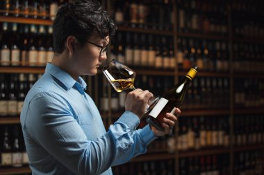 Açık mavi gömlekli şarap garsonu, dikkatle bir bardak şarap örneği alıyor, etiketini okumak için yanında tutuyor ve şarap raflarıyla dolu arka plandaki şişeleri okuyor.. 