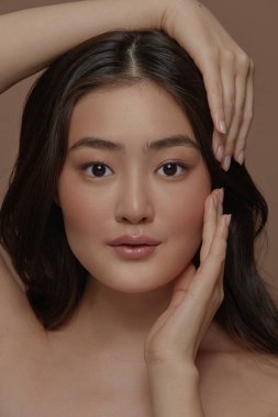 Uzun esmer, çekici, çekici bir Japon kadının portresinin dikey çekimi. Bej bir arka planda tek başına duran yüzüne dokunuyor. Cilt bakımı, kozmetoloji ve plastik